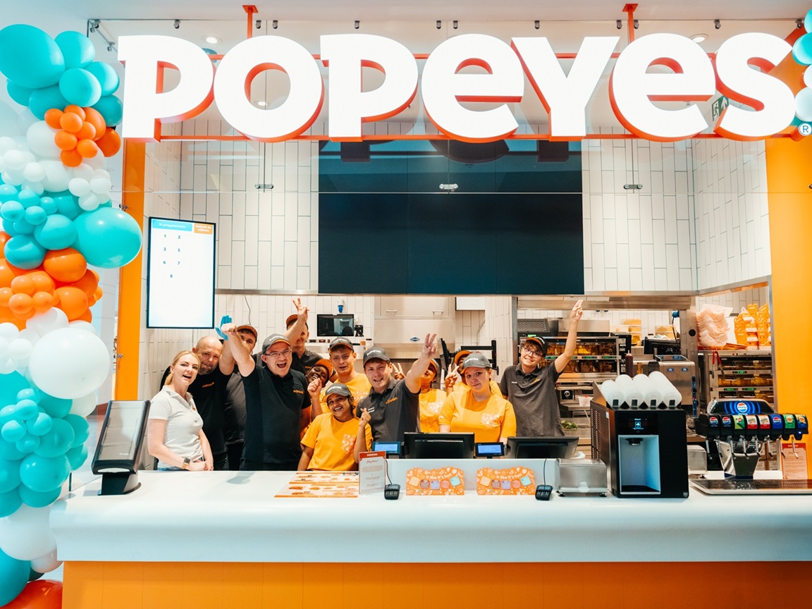 Grupa uśmiechniętych pracowników Popeyes w pomarańczowych i czarnych strojach pozuje za białą ladą restauracji z logo marki.