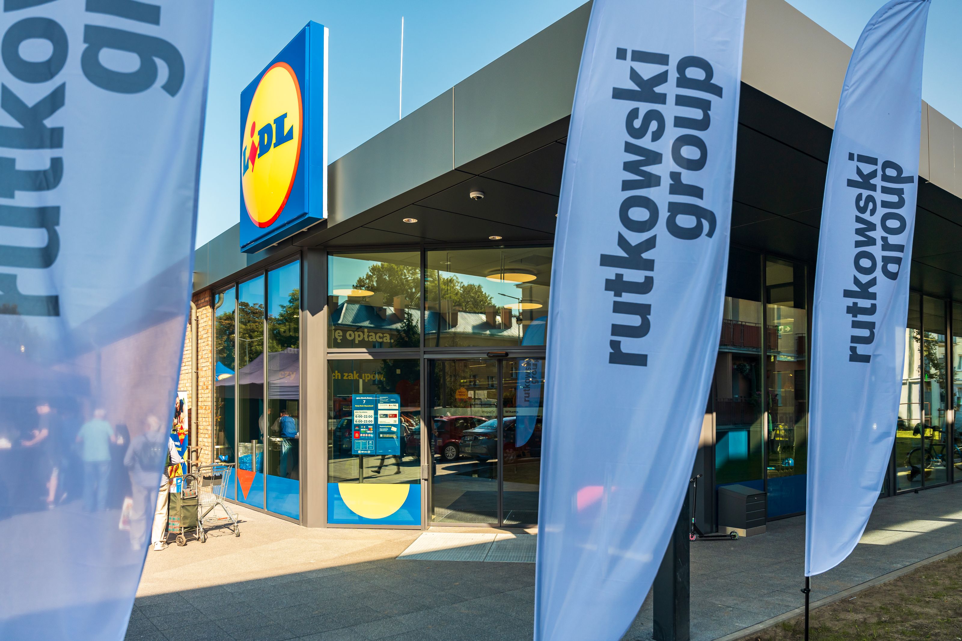 Nowoczesny budynek supermarketu Lidl z niebiesko-żółtym logo nad wejściem. Fasada z ciemnoszarych paneli i dużych witryn. Przed wejściem wysokie flagi reklamowe "rutkowski group". Klienci z wózkami zakupowymi na pierwszym planie.