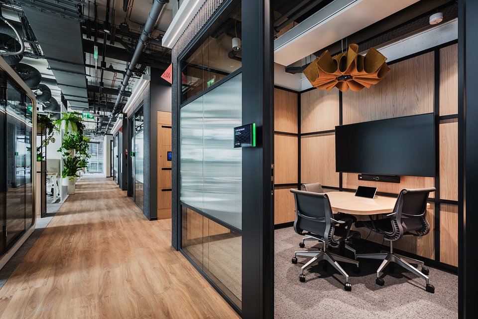 Nowoczesne wnętrze biurowe typu open space z przeszkloną salką spotkań, wyposażoną w ergonomiczne fotele i duży ekran ścienny.