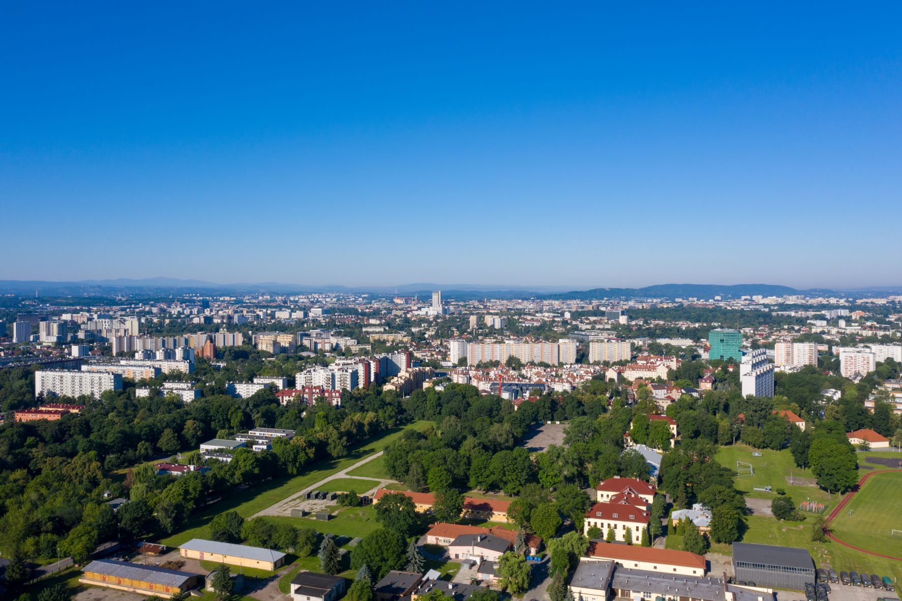 Panorama miasta z lotu ptaka ukazuje rozległą zabudowę mieszkaniową, liczne tereny zielone, niższe budynki, boiska oraz obszary magazynowe.