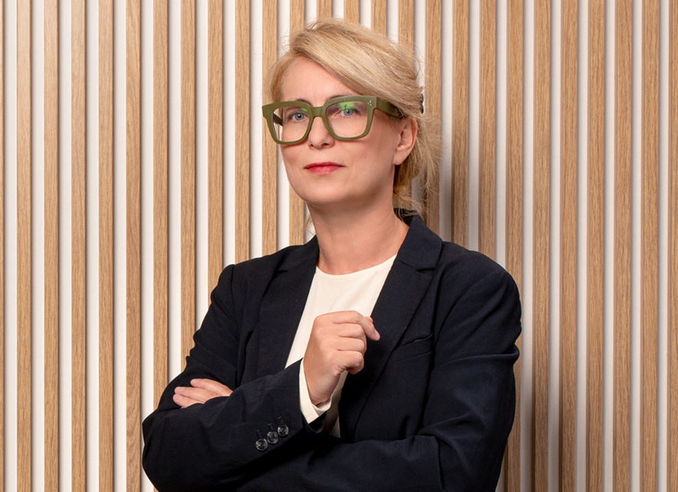 Portret Barbary Pryszcz: kobieta z blond włosami, w okularach oliwkowych, ciemnej marynarce, jasnej bluzce na tle drewnianych lamel.