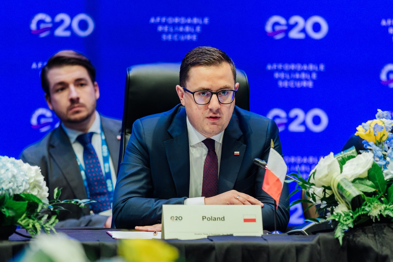 Mężczyzna w okularach, ubrany w ciemny garnitur i bordowy krawat, przemawia przy stole konferencyjnym.