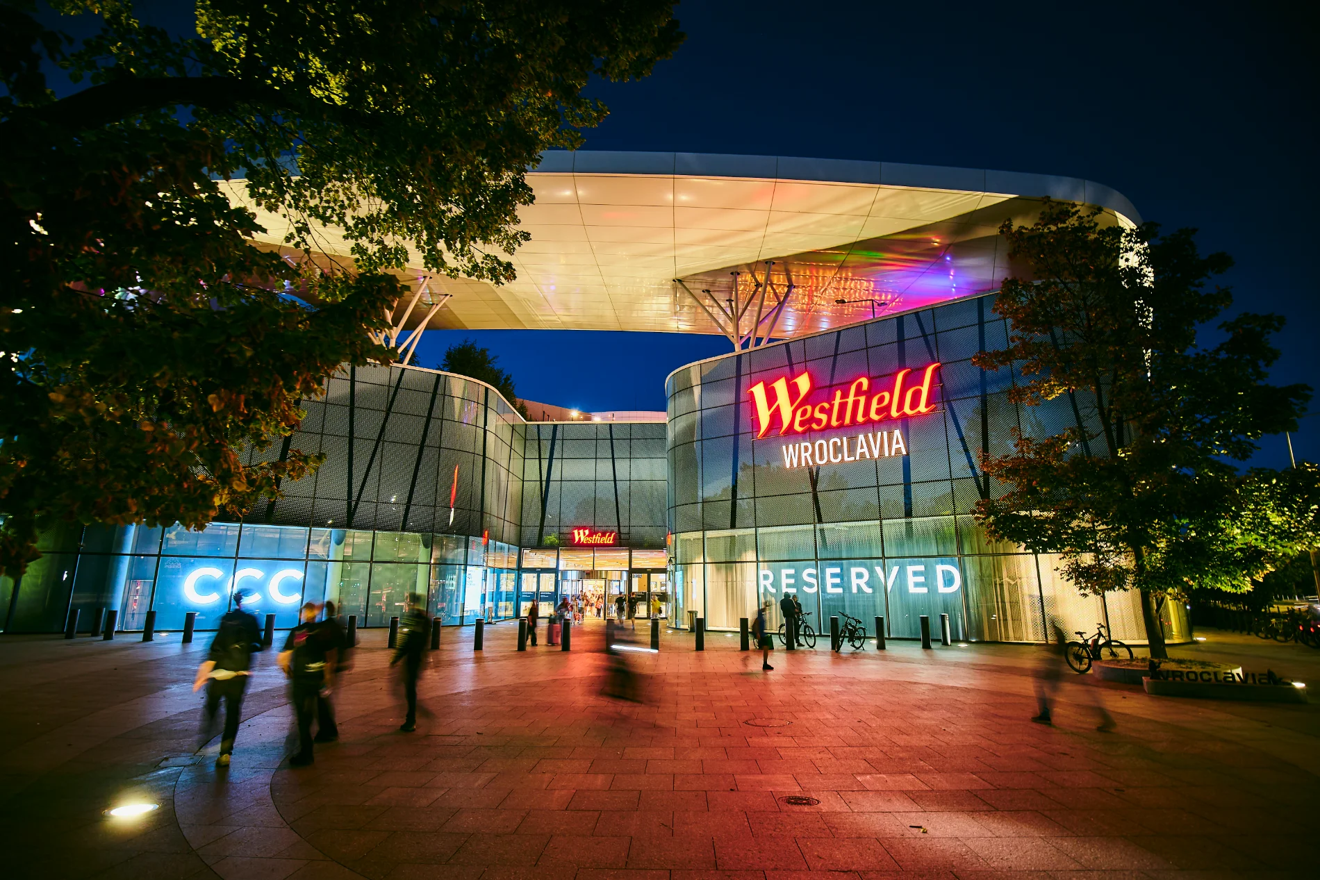 Oświetlona fasada Westfield Wroclavia o zmierzchu z widocznymi logo marek CCC i Reserved.