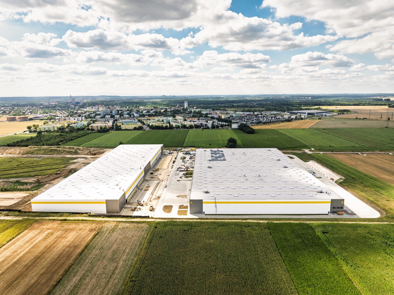 Lotniczy widok na dwa nowoczesne centra logistyczne z jasnymi dachami i żółtymi pasami, otoczone polami uprawnymi, z panoramą miasta w oddali.