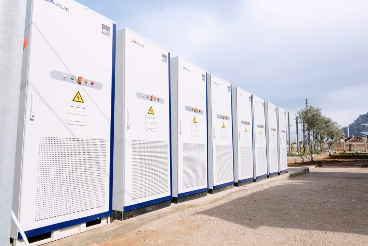 Szereg białych, wysokich szaf technologicznych marki SolarEdge stoi na betonowym podłożu. Urządzenia mają niebieskie wykończenia, panele kontrolne z przyciskami i żółte znaki ostrzegawcze z błyskawicą. W tle widać drzewa oliwne, niską zabudowę i góry pod zachmurzonym niebem.