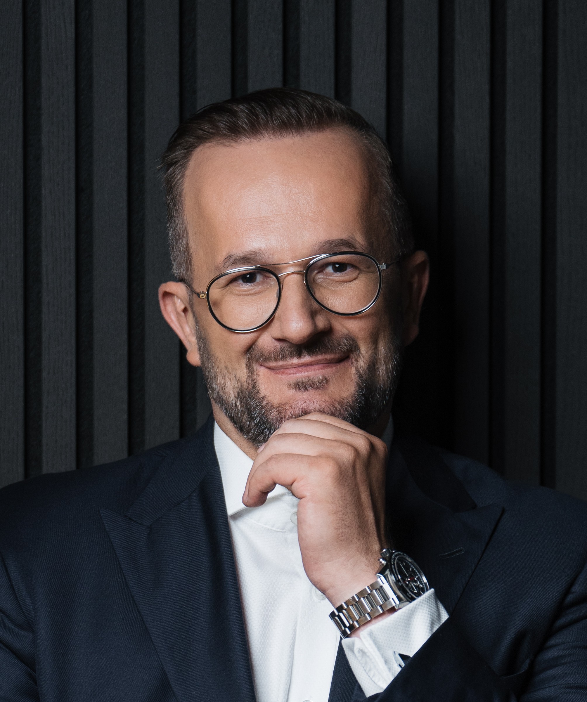 Mężczyzna z krótkim zarostem i zaczesanymi włosami w okularach i marynarce, uśmiecha się, opierając podbródek na dłoni, na tle czarnych lamel.