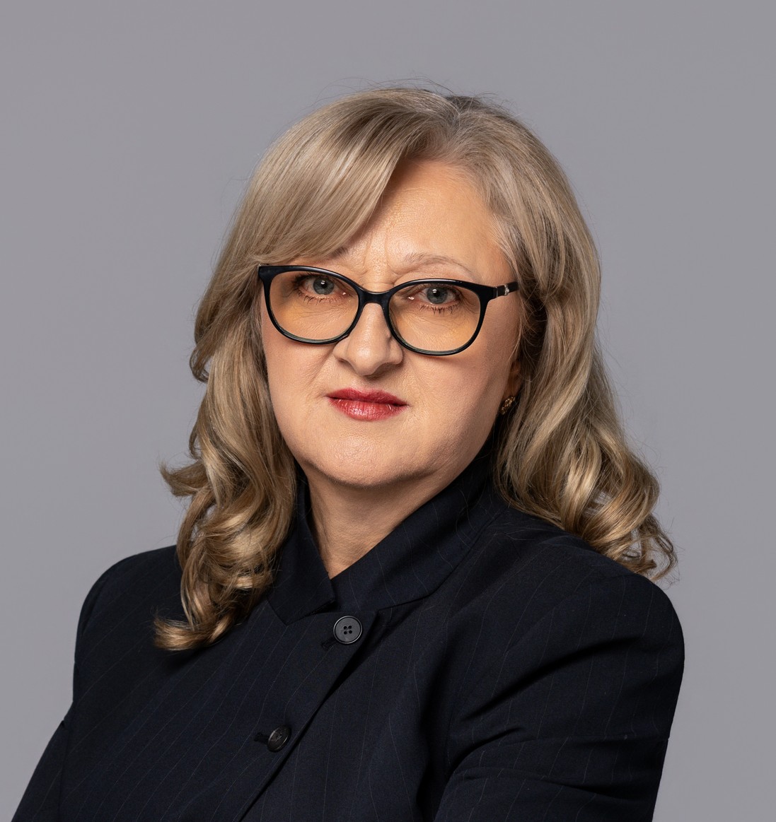 Portret dojrzałej kobiety o blond włosach średniej długości w okularach i ciemnej marynarce na szarym tle.