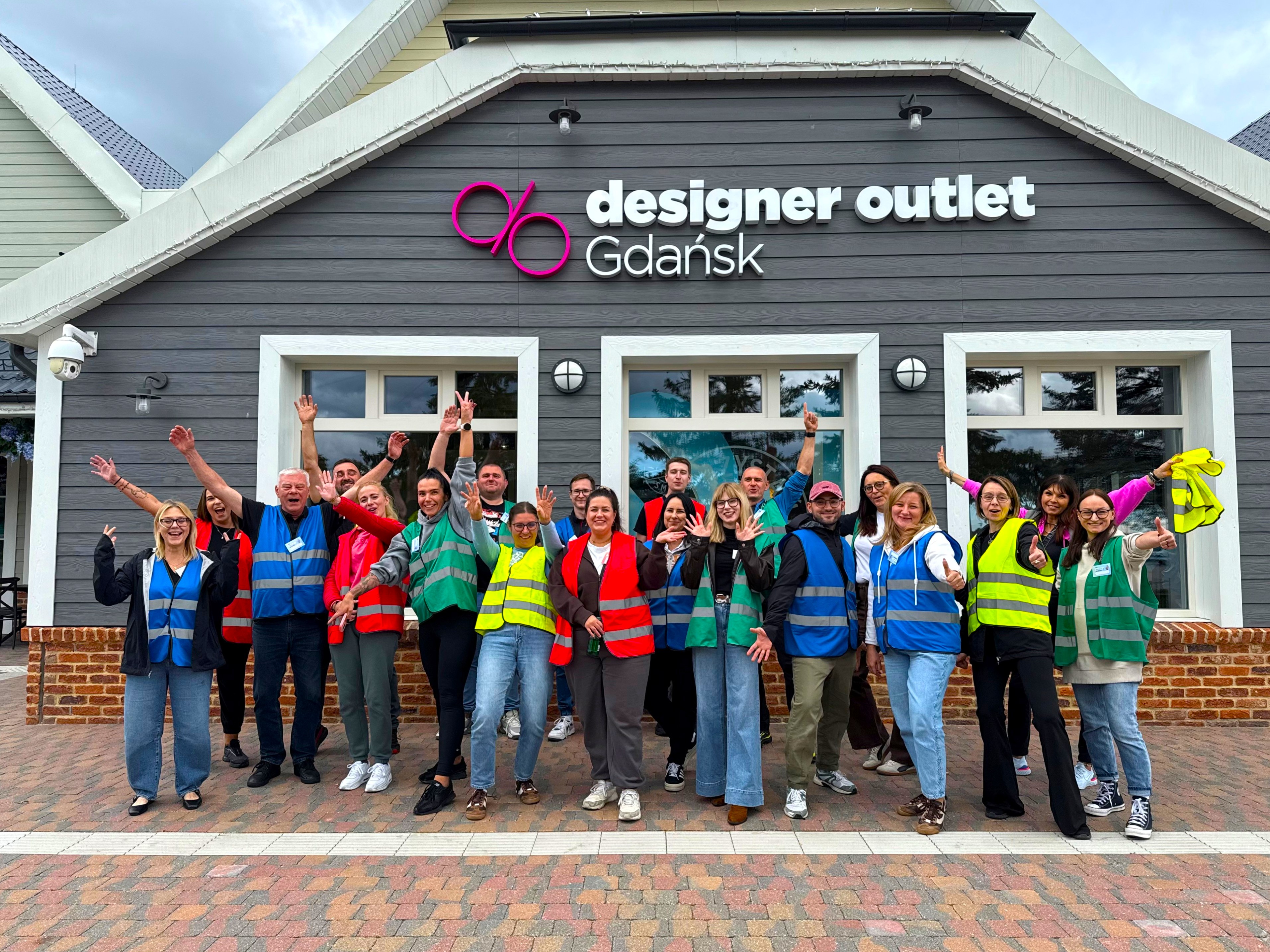 Uśmiechnięta grupa kilkunastu osób w różnym wieku pozuje do zdjęcia przed budynkiem Designer Outlet Gdańsk w kolorowych kamizelkach odblaskowych.