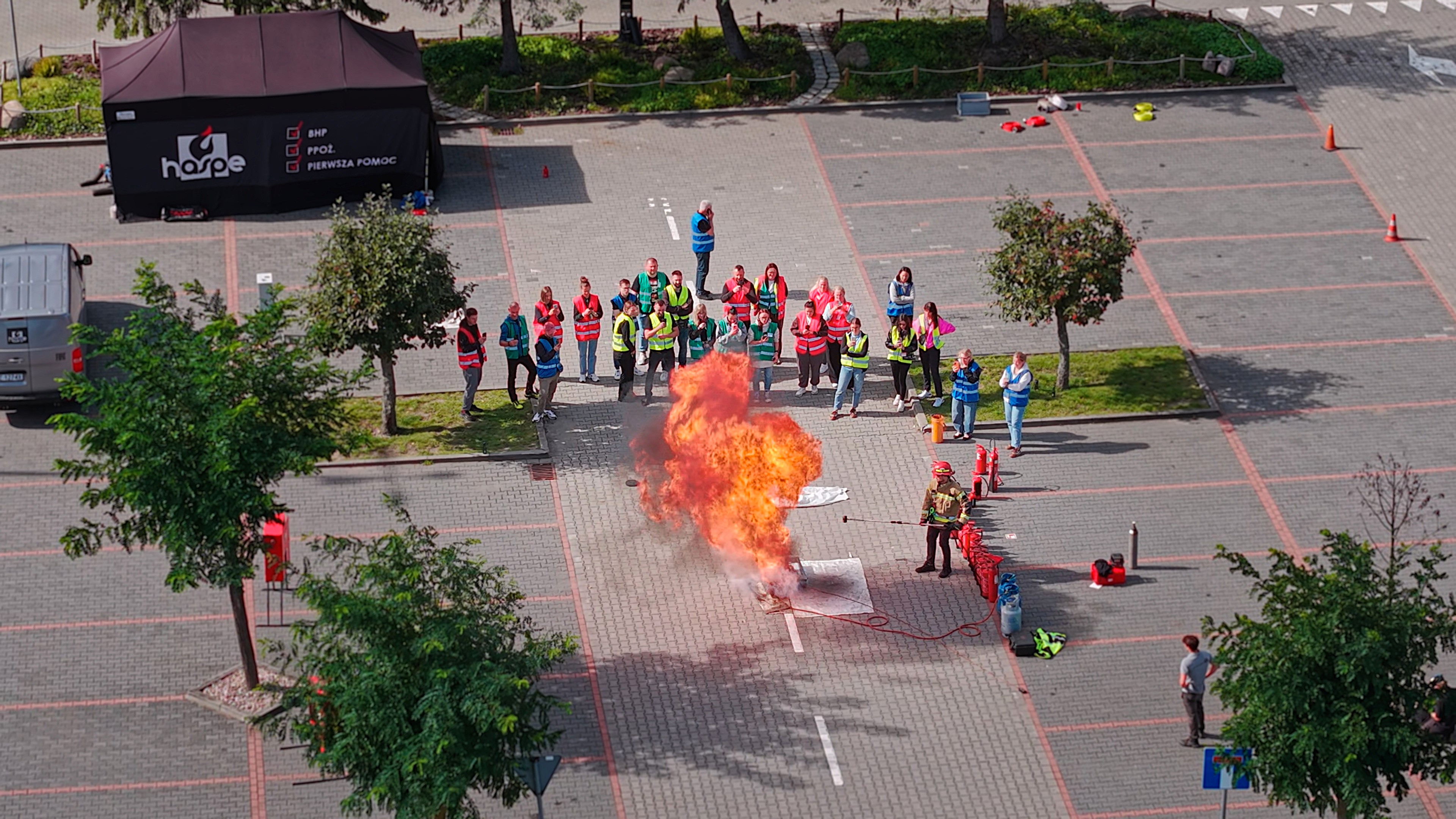 Strażak gasi płomienie na wybrukowanym placu, a grupa osób w kamizelkach obserwuje zdarzenie z bezpiecznej odległości.