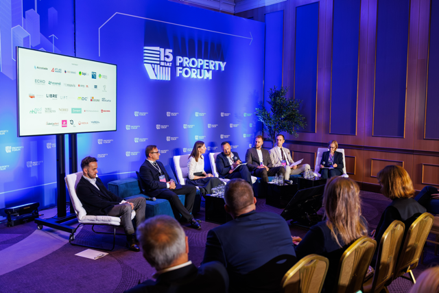 Grupa prelegentów w nowoczesnych fotelach na scenie Property Forum przed niebieskim tłem z logotypami.