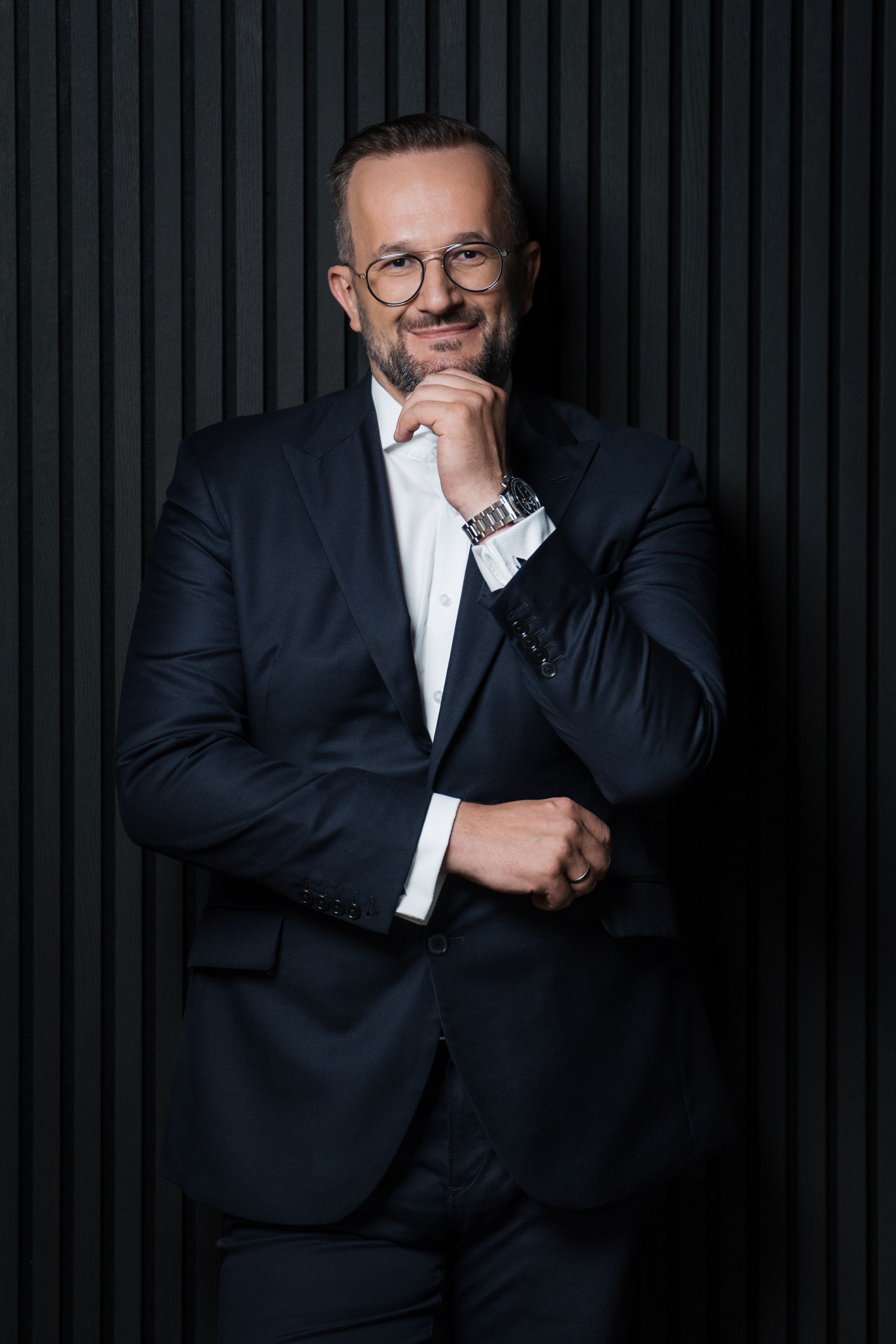 Mężczyzna w średnim wieku, z krótkim zarostem i w okularach, pozuje na tle ściany wyłożonej czarnymi lamelami, ubrany w elegancki, ciemnogranatowy garnitur.