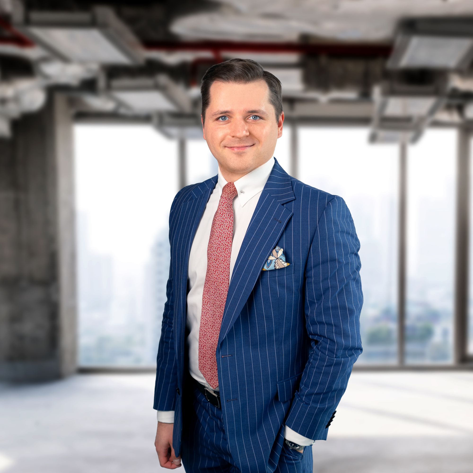 Mężczyzna w średnim wieku pozuje w eleganckim, niebieskim garniturze w białe prążki, białej koszuli i czerwonym krawacie. Stoi z ręką w kieszeni na tle rozmytego, jasnego wnętrza biurowca.