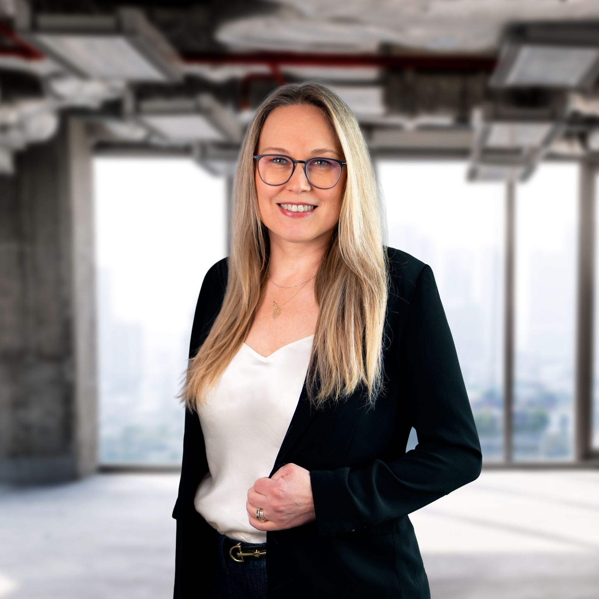 Uśmiechnięta kobieta o długich blond włosach w okularach, ubrana w czarną marynarkę i białą bluzkę, stoi w nowoczesnym, przeszklonym pomieszczeniu z panoramą miasta w tle.
