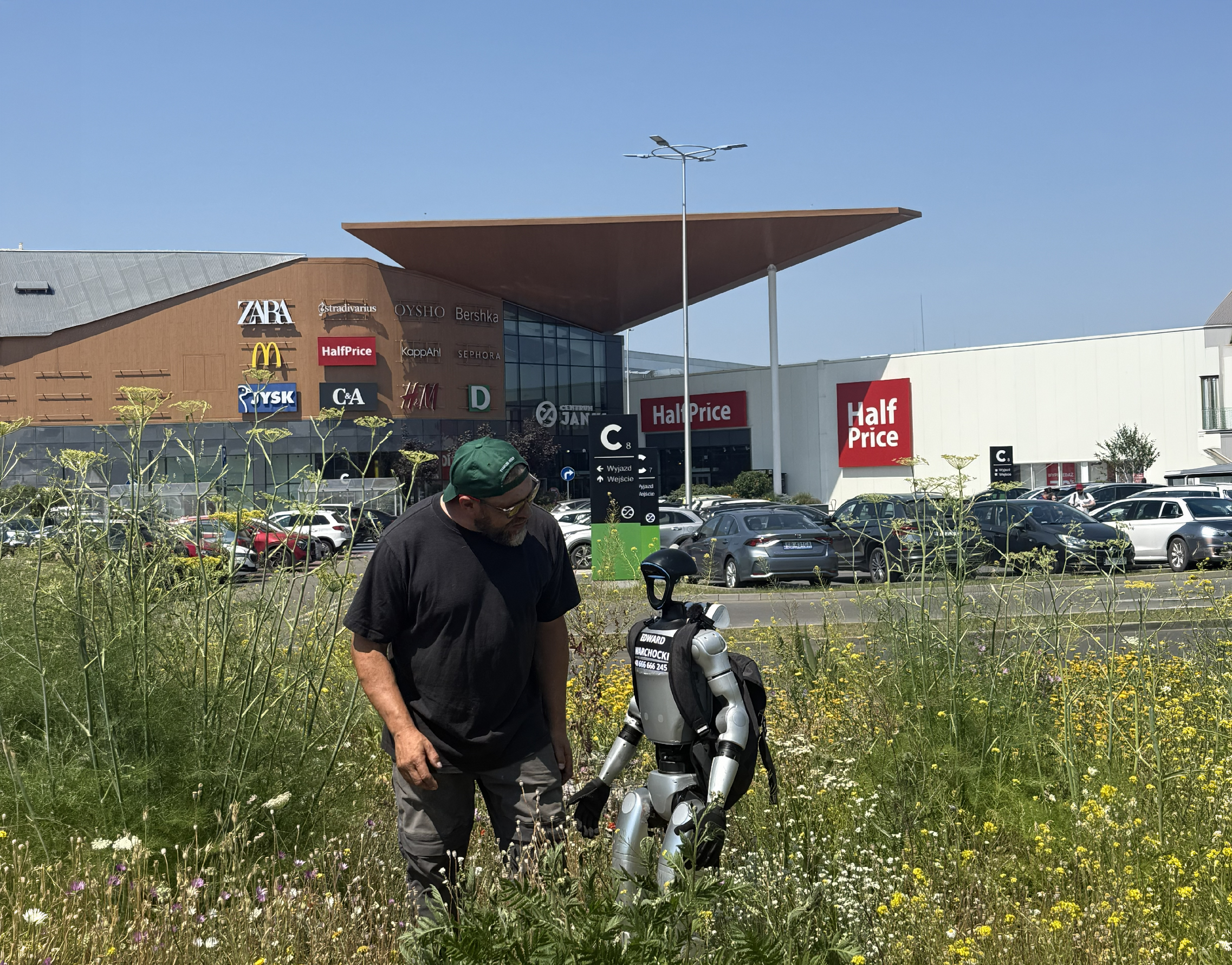 Mężczyzna w zielonej czapce stoi na łące kwietnej obok srebrno-czarnego humanoidalnego robota z plecakiem.