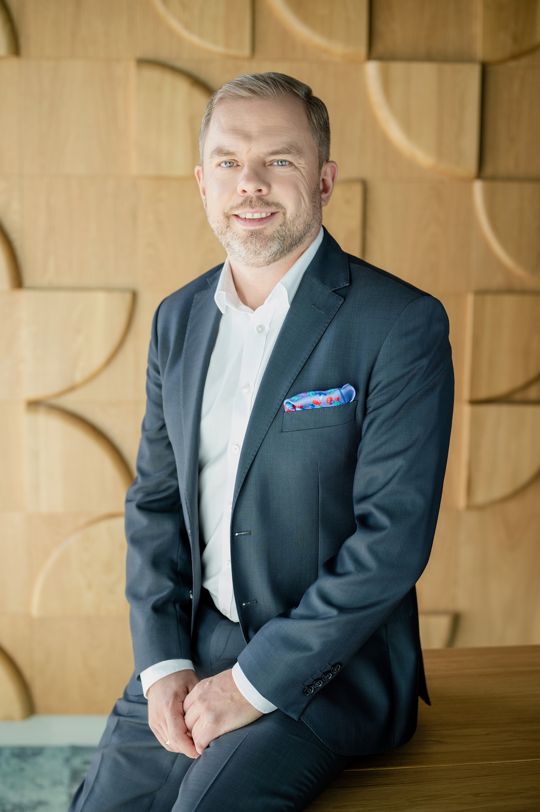 Marcin Sulewski, uśmiechnięty mężczyzna z krótkim zarostem, w ciemnoszarej marynarce, białej koszuli i wzorzystej poszetce, siedzi na drewnianym blacie.