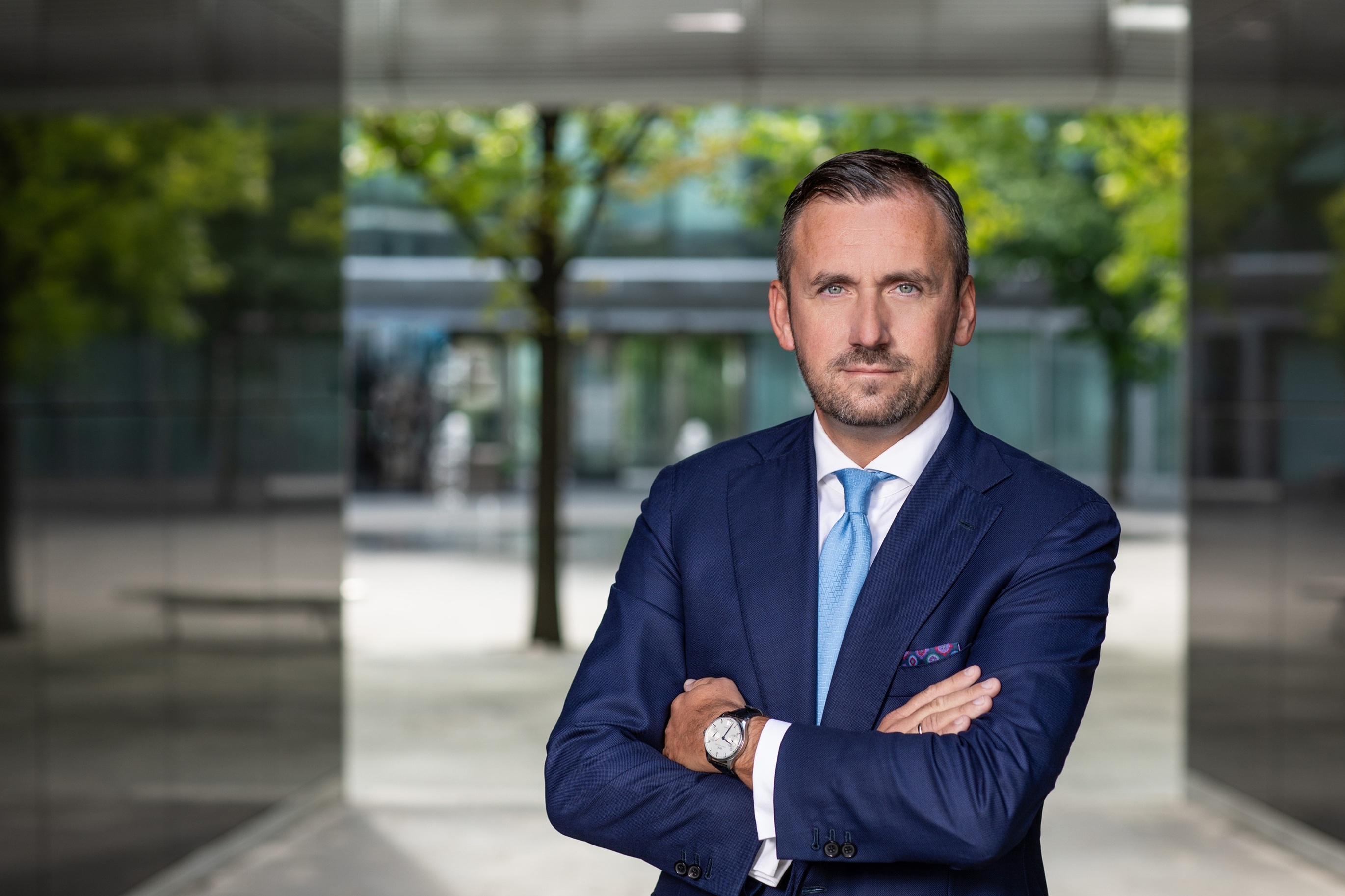 Piotr Mirowski, mężczyzna w garniturze z niebieskimi oczami i krótkim zarostem, pozuje z założonymi rękami na tle nowoczesnej architektury.