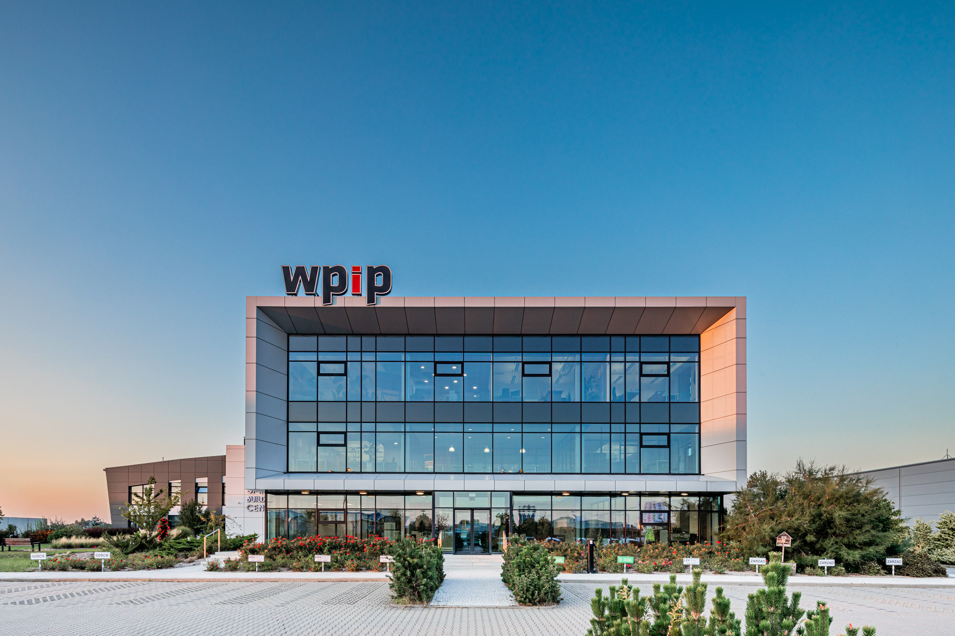Nowoczesny trzykondygnacyjny biurowiec o przeszklonej fasadzie z logotypem „wpip” na dachu, o zmierzchu.