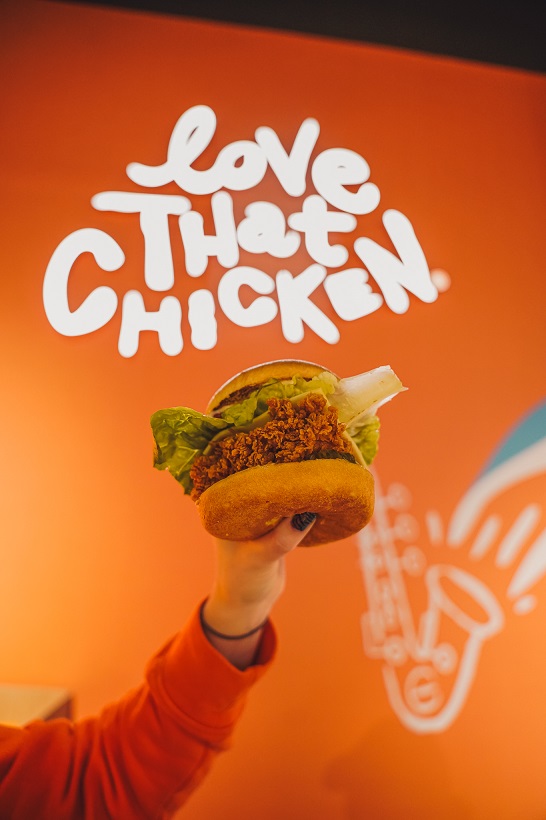 Dłoń w pomarańczowym rękawie unosi kanapkę z chrupiącym kurczakiem i sałatą na tle pomarańczowej ściany z białym napisem „Love THAT CHICKEN.” i grafiką saksofonu.