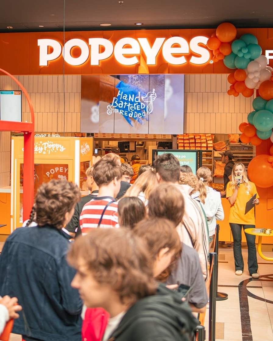 Długa kolejka młodych ludzi przed pomarańczową witryną restauracji Popeyes z dużym, świecącym napisem, pracownicą z mikrofonem i kolorowymi balonami.
