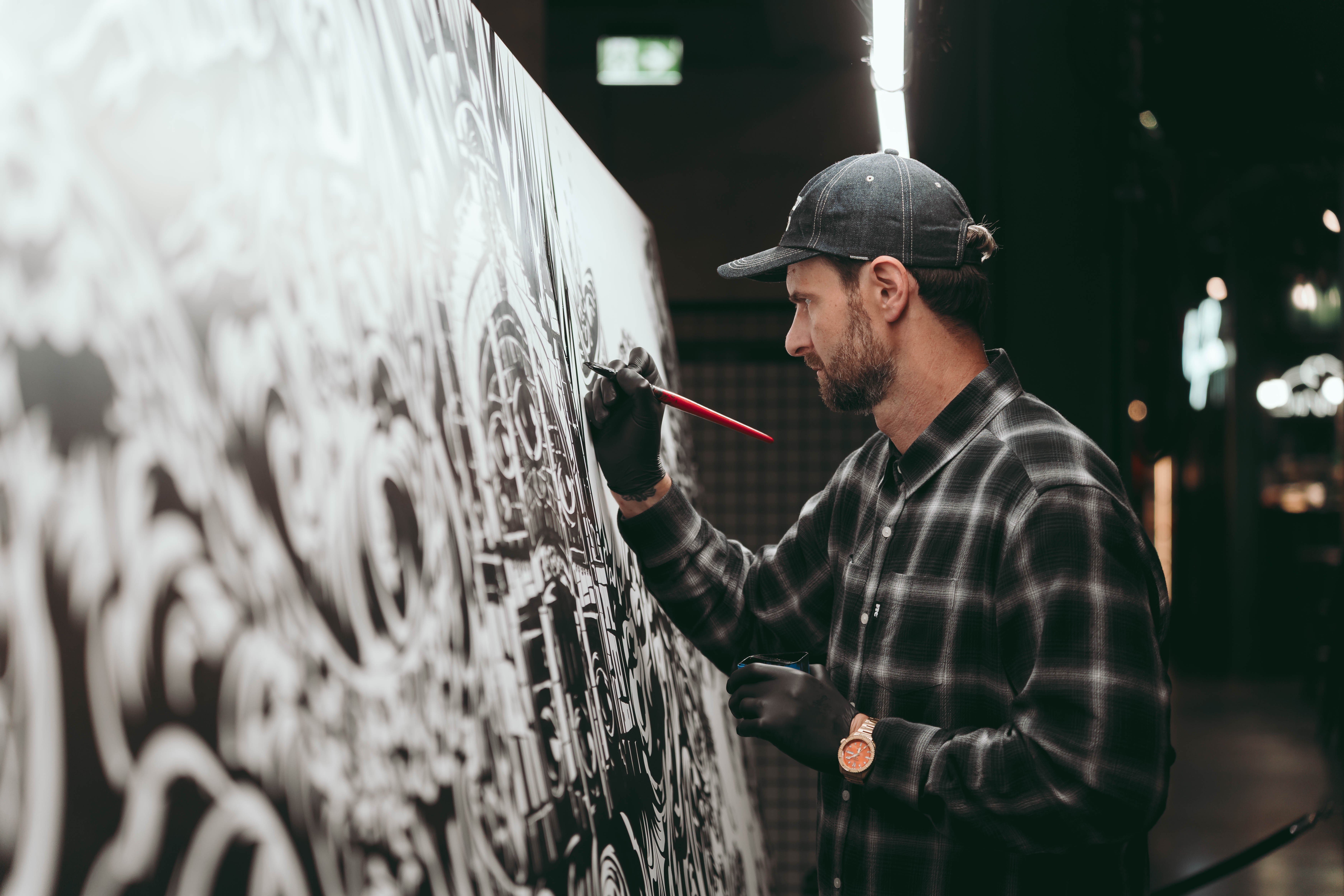 Mężczyzna w czapce z daszkiem i flanelowej koszuli maluje pędzlem czarno-biały mural na dużej, pionowej płaszczyźnie, z gęstymi, abstrakcyjnymi wzorami przypominającymi architekturę.
