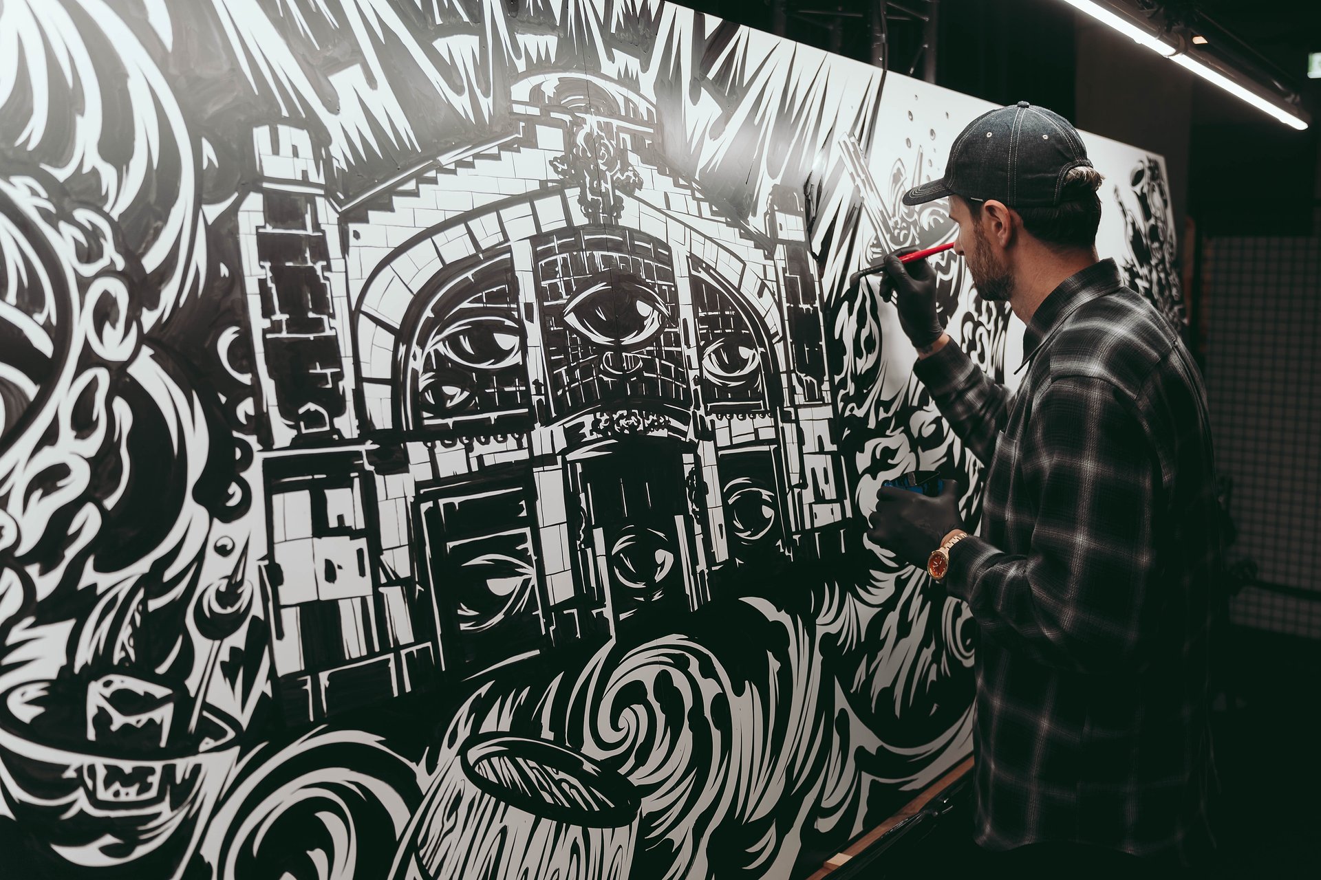 Mężczyzna w czapce i koszuli w kratę maluje mural czarną farbą na dużym, białym panelu, przedstawiającym budynek w stylu klasycystycznym z surrealistycznymi, dynamicznymi wzorami i motywem oczu.