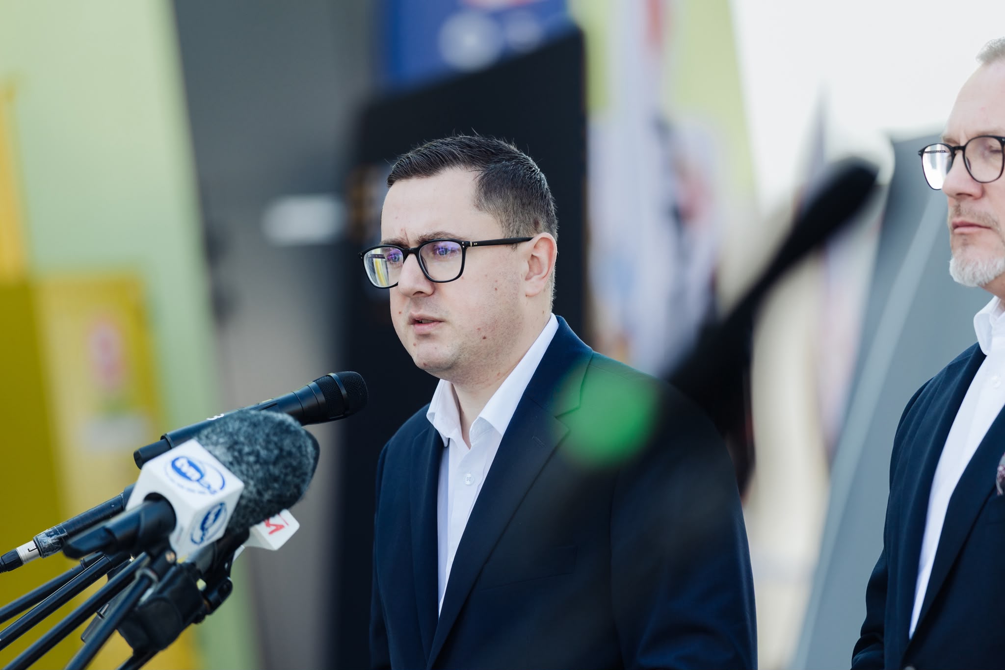Mężczyzna w ciemnym garniturze i okularach przemawia do licznych mikrofonów stacji telewizyjnych.