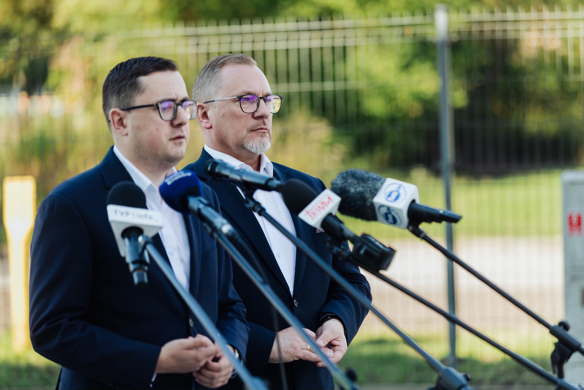 Dwaj mężczyźni w ciemnych garniturach i okularach stoją przed mikrofonami na konferencji prasowej.