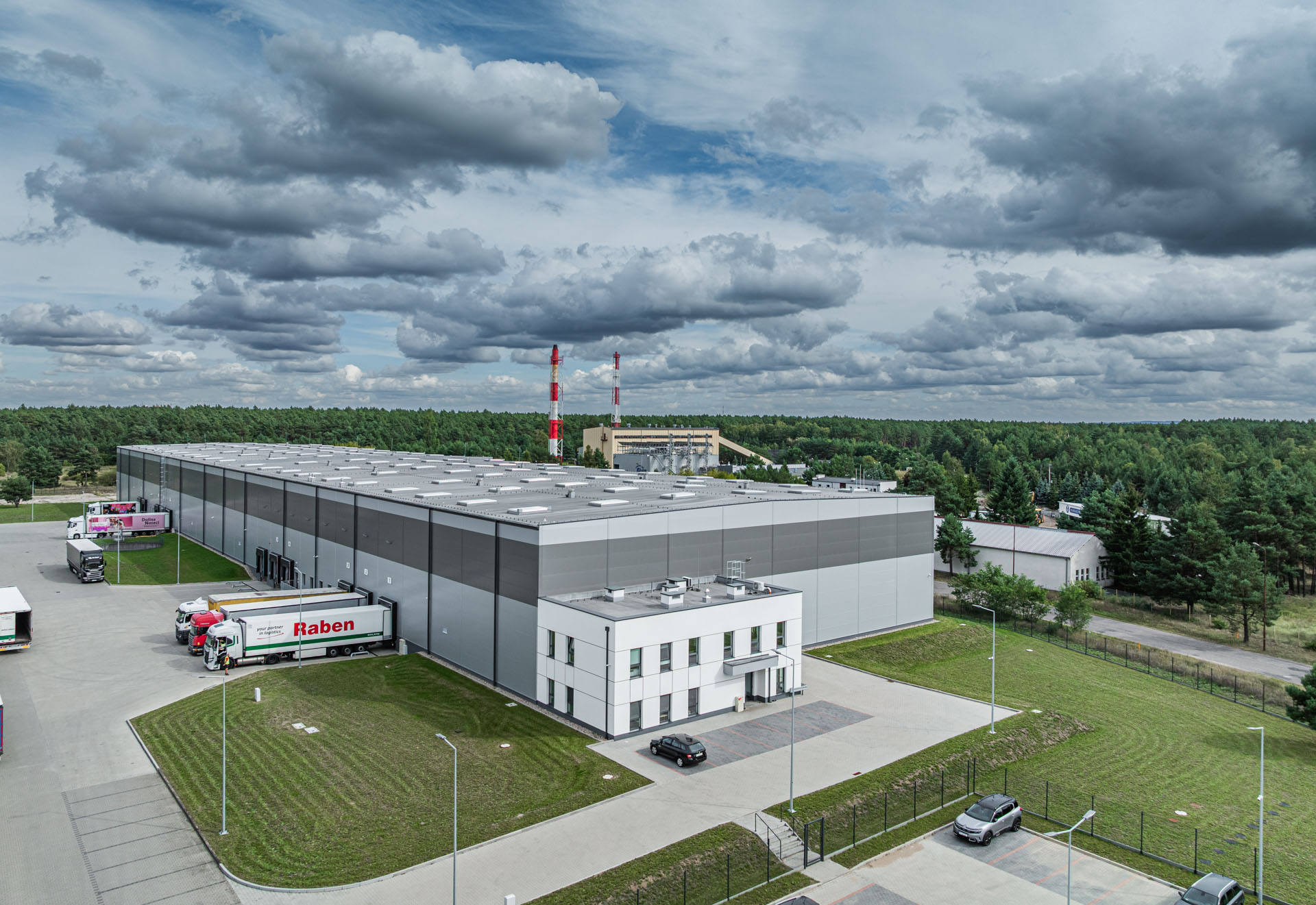 Nowoczesna hala magazynowa z szarą elewacją, przylegającym białym biurowcem, samochodami ciężarowymi przy dokach przeładunkowych oraz zielonymi trawnikami.