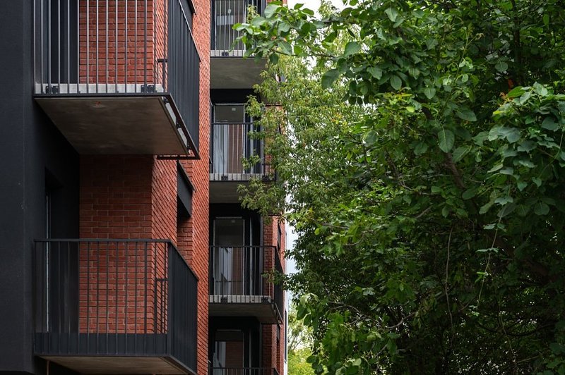 Nowoczesny budynek wielorodzinny o elewacji z czerwonej cegły i ciemnego tynku. Ma liczne balkony z czarnymi, prostymi balustradami z pionowych prętów. Budynek sąsiaduje z gęstą, zieloną roślinnością, która częściowo przesłania fasadę.