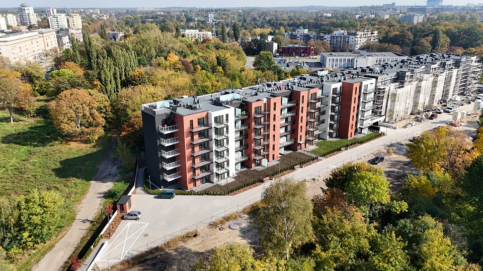 Widok z lotu ptaka przedstawia nowoczesny, wielorodzinny budynek mieszkalny z czerwoną cegłą, białym tynkiem i ciemnoszarymi panelami, otoczony gęstą roślinnością. W tle panorama miasta, a na pierwszym planie widoczne są rusztowania budowlane.