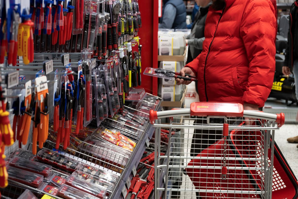 Mężczyzna w czerwonej kurtce puchowej ogląda zapakowane kombinerki przy regale z narzędziami ręcznymi, obok metalowego wózka zakupowego.