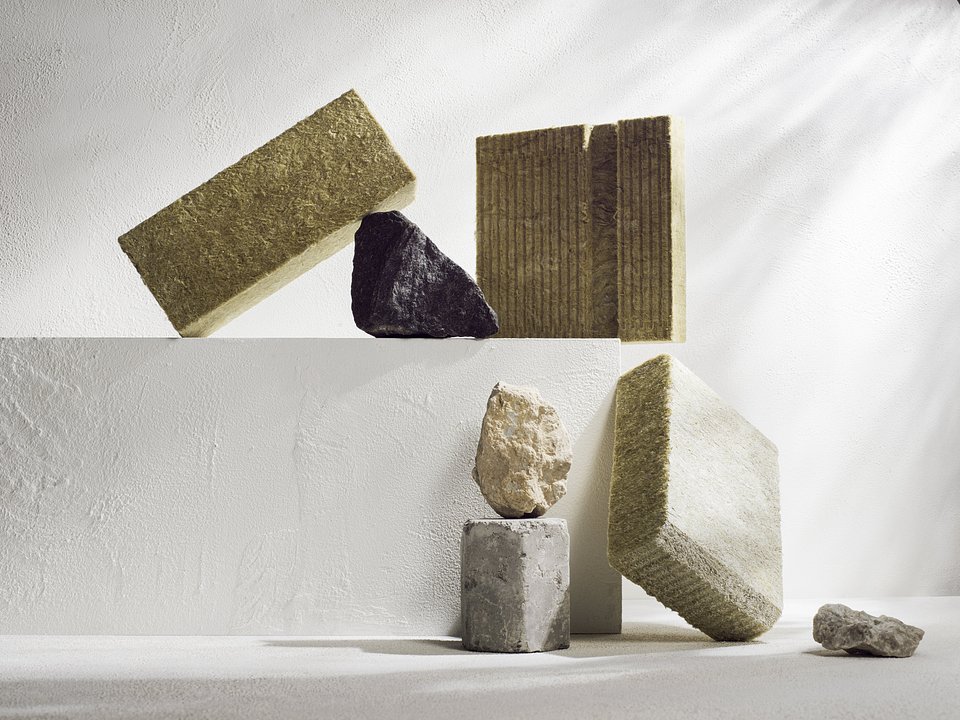 Bloki zielonej i beżowej wełny mineralnej ułożone z kamieniami oraz betonowym postumentem na tle białej ściany w minimalistycznym studiu, z delikatnymi cieniami.