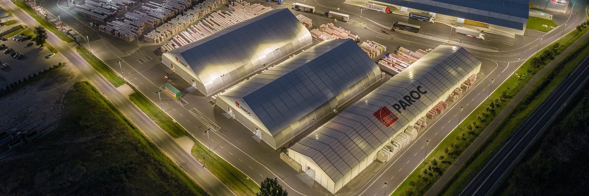 Lotnicze ujęcie kompleksu trzech jasnoszarych hal namiotowych z logo Paroc i Owens Corning, otoczonych placem składowym, drogami dojazdowymi i ciężarówkami, oświetlonych w zmierzchu.