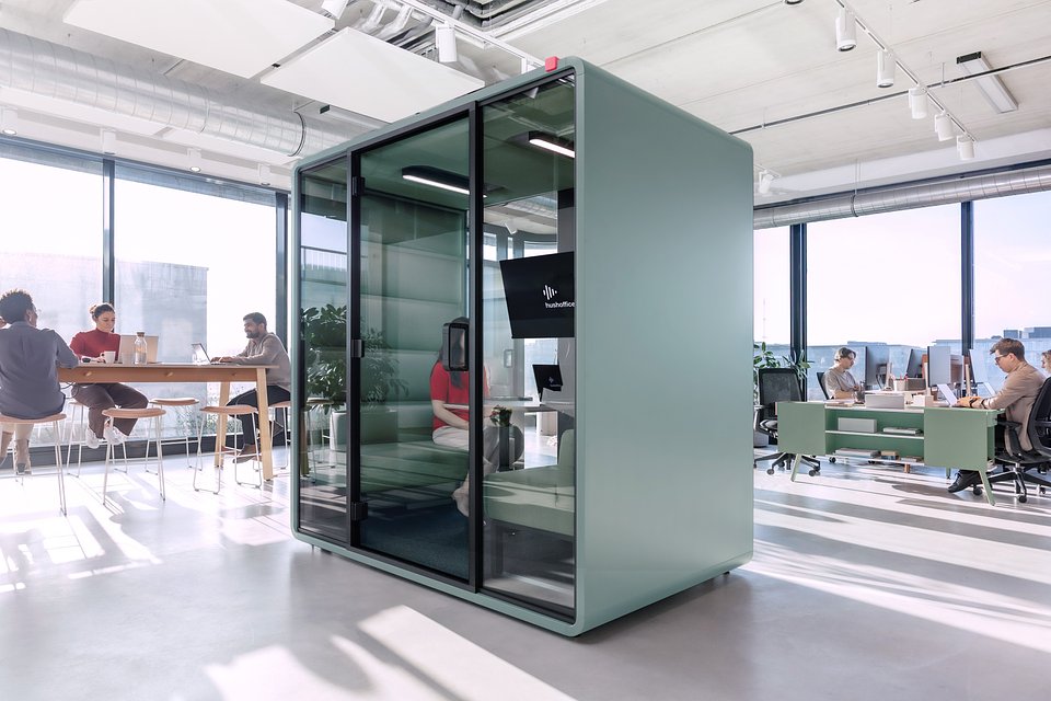 Nowoczesna, przeszklona kabina biurowa typu „acoustic pod” w kolorze szałwiowym, stojąca w jasnym biurze open space.