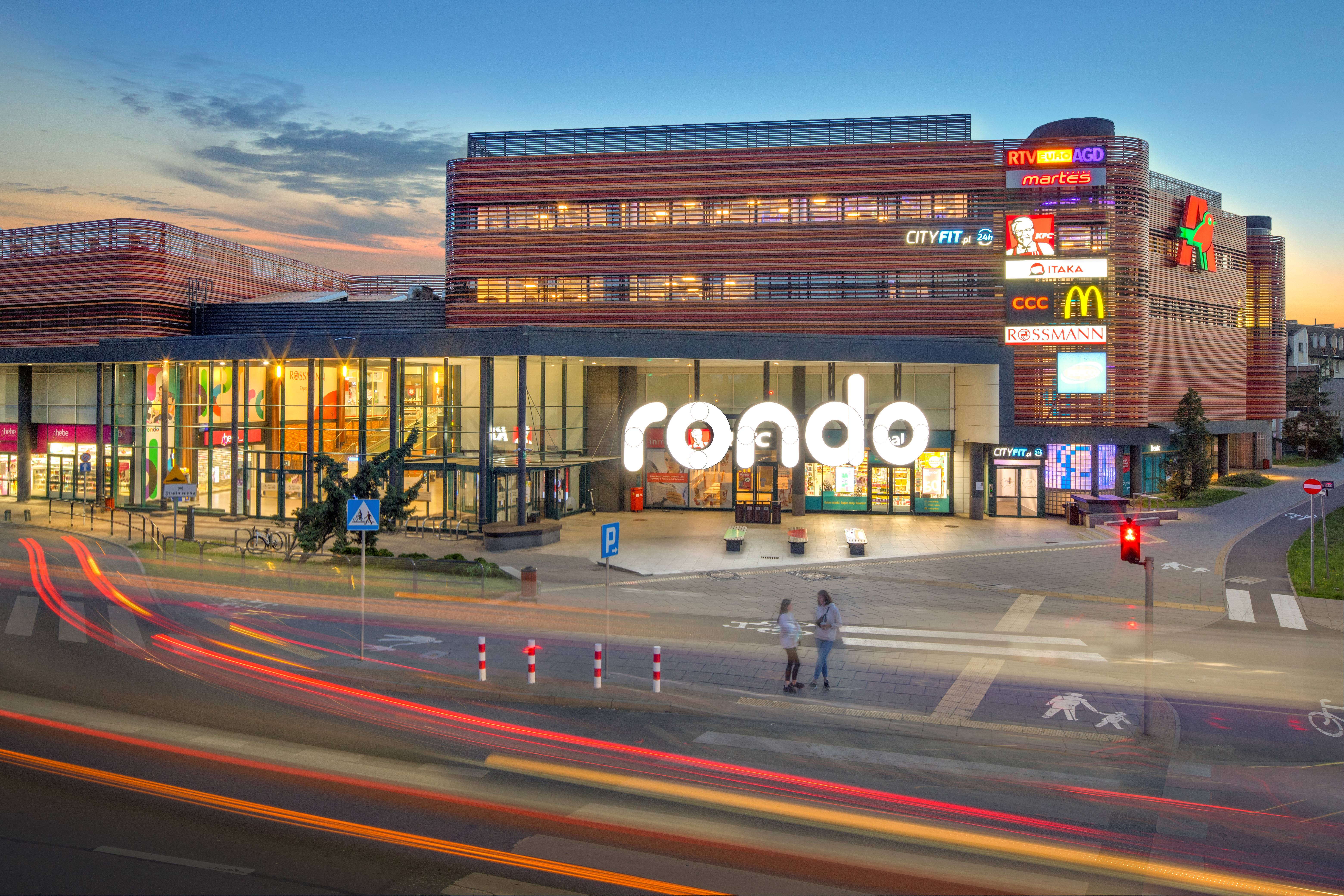 Wielokondygnacyjne centrum handlowe Rondo z pomarańczowo-brązową elewacją, przeszklonymi witrynami, licznymi logotypami marek i oświetlonym placem.