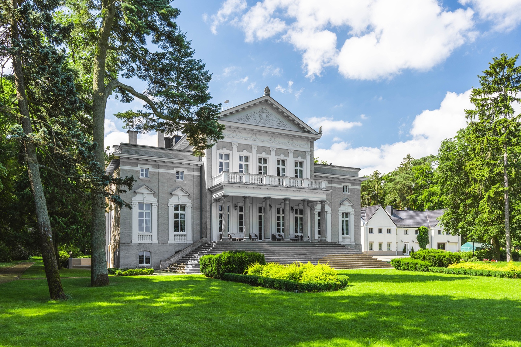 Klasycystyczny, szary pałac z białymi zdobieniami i kolumnowym portykiem, otoczony zielonym ogrodem.