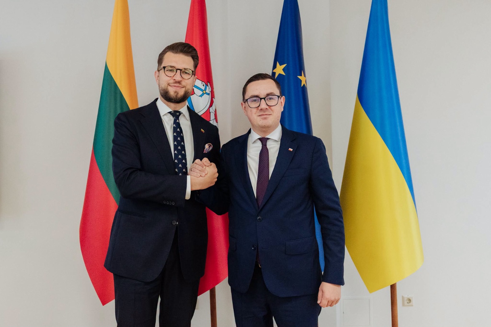 Dwaj mężczyźni w garniturach podają sobie ręce na tle flag państwowych i Unii Europejskiej.