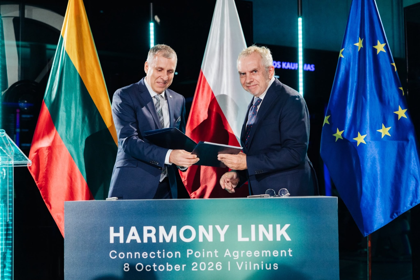 Dwaj mężczyźni w garniturach trzymają otwarte teczki za mównicą Harmony Link, Wilno 2026.