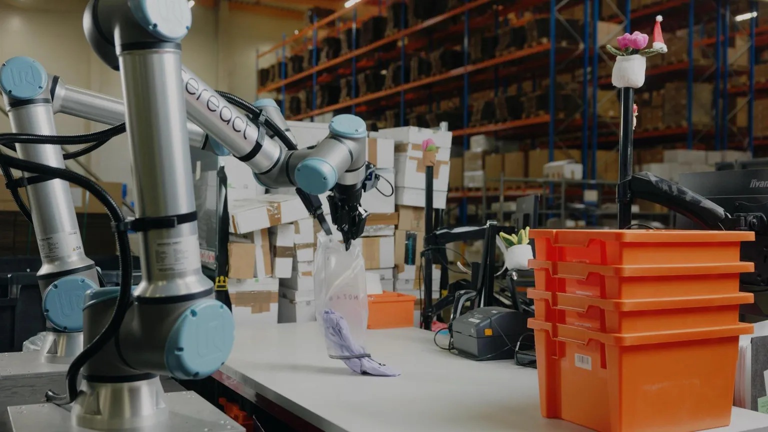 Srebrno-niebieskie ramię robota przemysłowego chwyta przezroczystą torbę z fioletowym materiałem nad białym blatem roboczym.