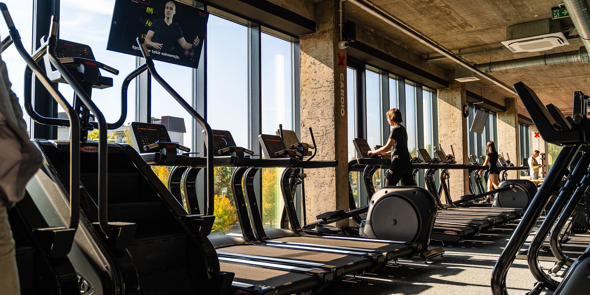 Nowoczesna sala fitness z rzędami czarnych bieżni i maszyn eliptycznych ustawionych wzdłuż przeszklonej ściany, przez którą wpada jasne światło słoneczne. W głębi widoczne są sylwetki ćwiczących osób, a wnętrze charakteryzuje się industrialnym stylem z surowym, betonowym sufitem i widocznymi instalacjami. Pod sufitem zamontowany jest ekran telewizyjny z wyświetlanym instruktorem, a całość utrzymana jest w ciemnej kolorystyce kontrastującej z widokiem jesiennych drzew za oknem.