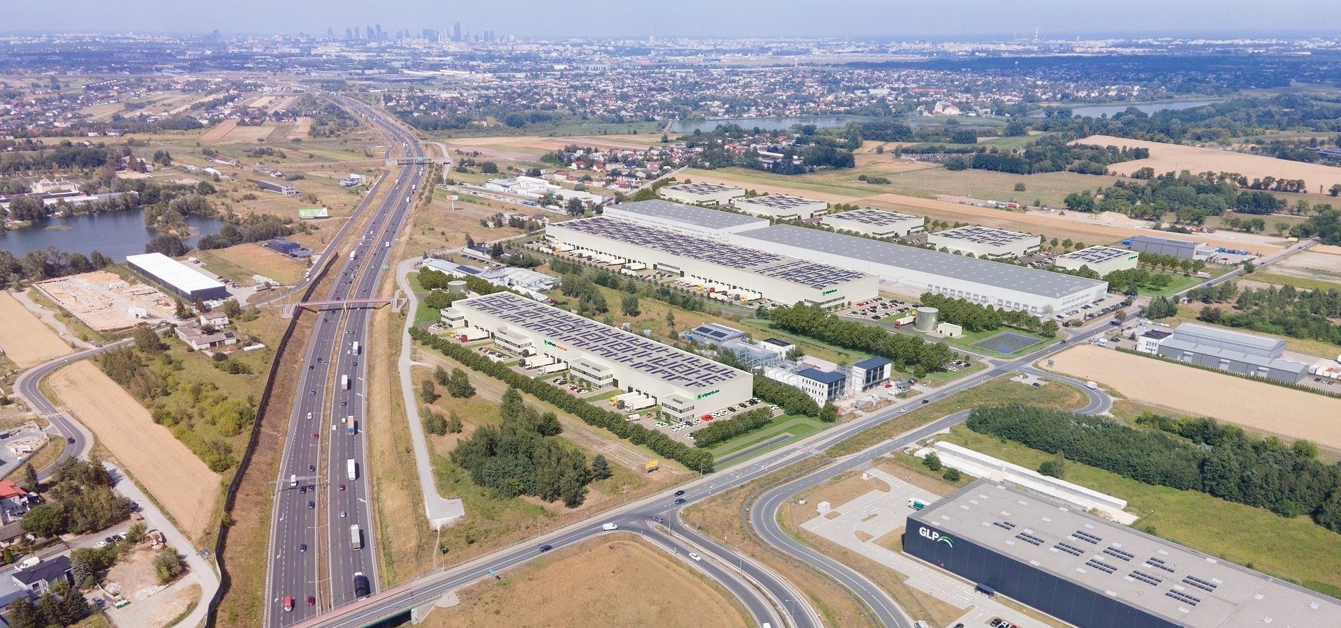 Widok lotniczy kompleksu hal logistycznych i magazynowych z jasnoszarymi elewacjami, zlokalizowanego przy autostradzie.
