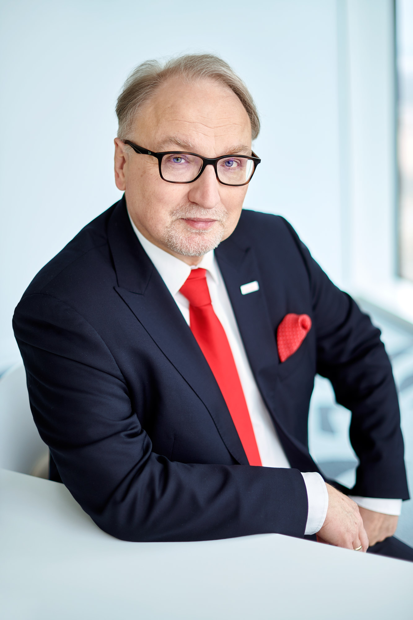 Portret dojrzałego mężczyzny w okularach, w ciemnogranatowym garniturze, białej koszuli i intensywnie czerwonym krawacie. Ma krótkie, siwiejące włosy i przystrzyżoną brodę. Siedzi pochylony, z ręką na białym blacie.