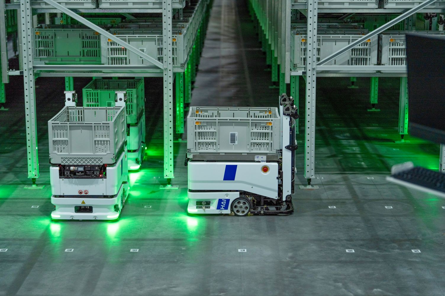 W nowoczesnym centrum logistycznym białe roboty autonomiczne z zielonym podświetleniem transportują szare pojemniki pomiędzy wysokimi regałami magazynowymi, w surowej hali z betonową posadzką.