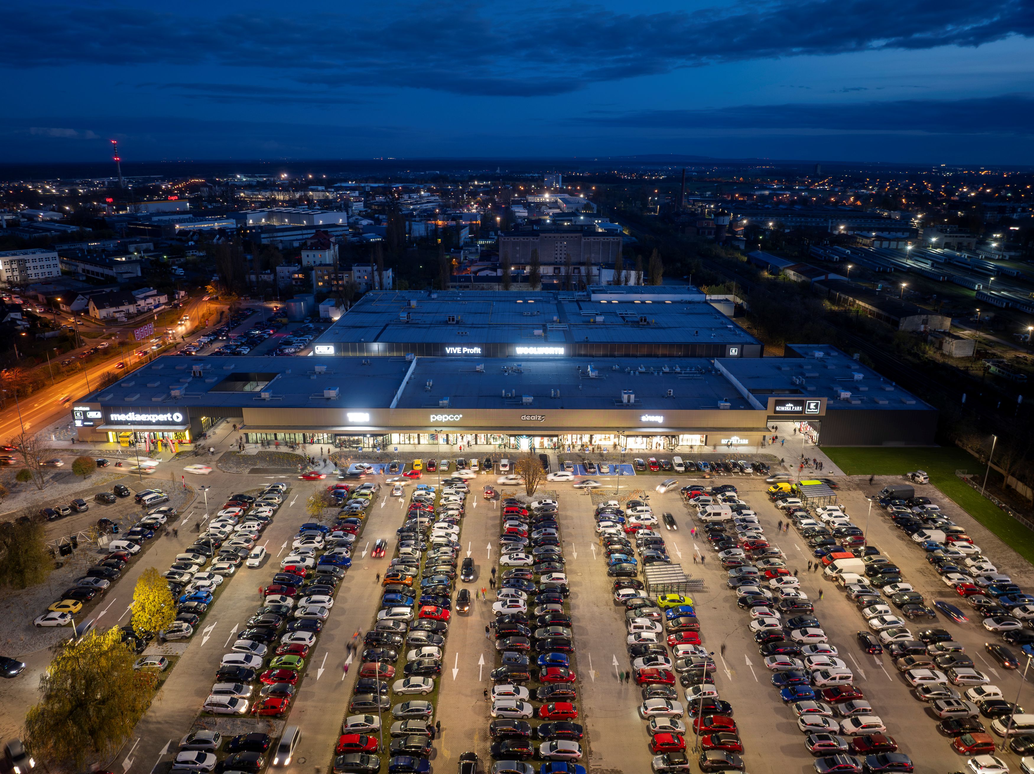 Widok z lotu ptaka na oświetlony park handlowy z dużym parkingiem o zmierzchu.