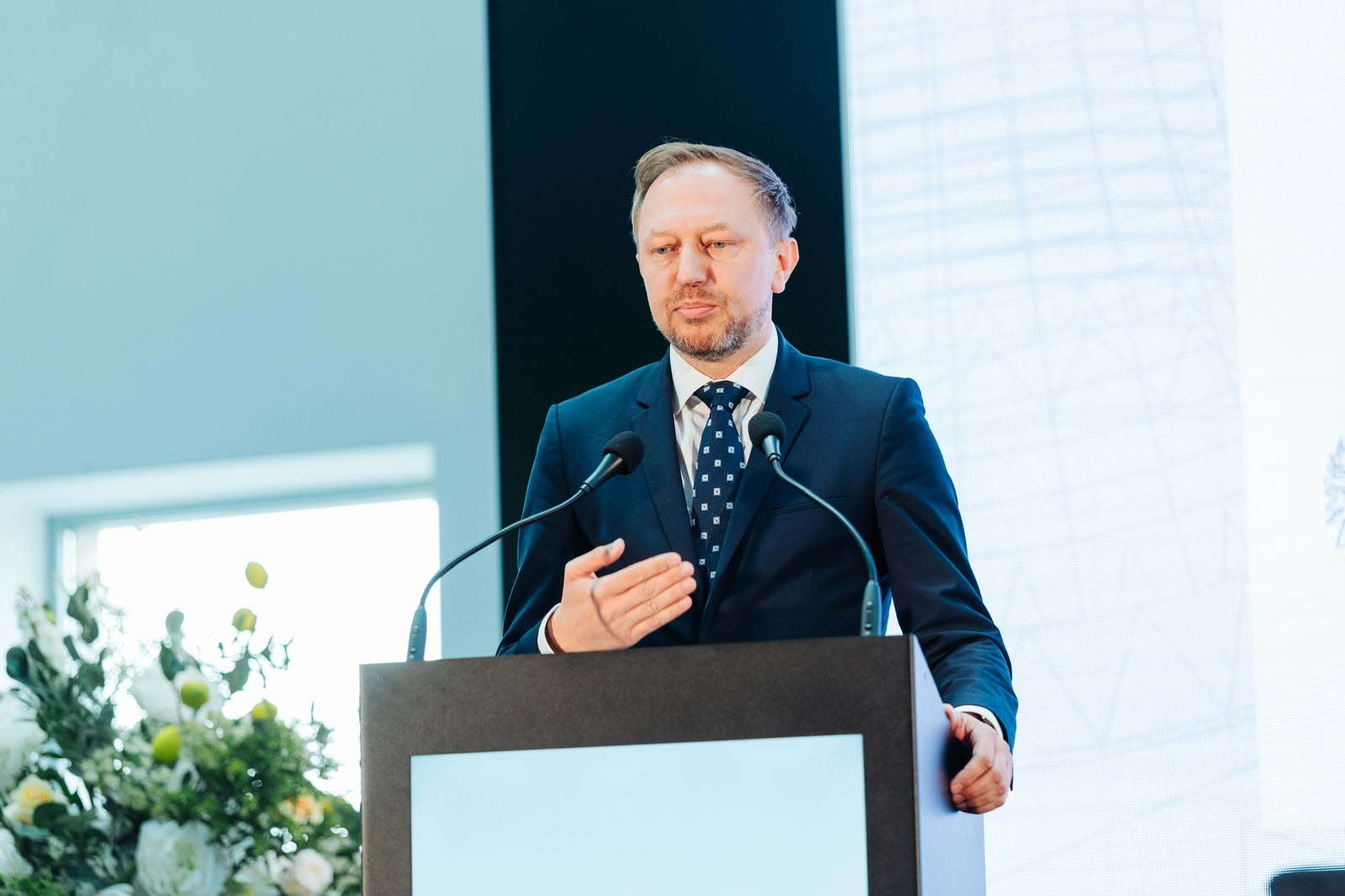 Mężczyzna w średnim wieku z krótkim zarostem przemawia za czarną mównicą z wbudowanym ekranem. W tle duży, jasny ekran oraz kompozycja z białych i żółtych kwiatów.