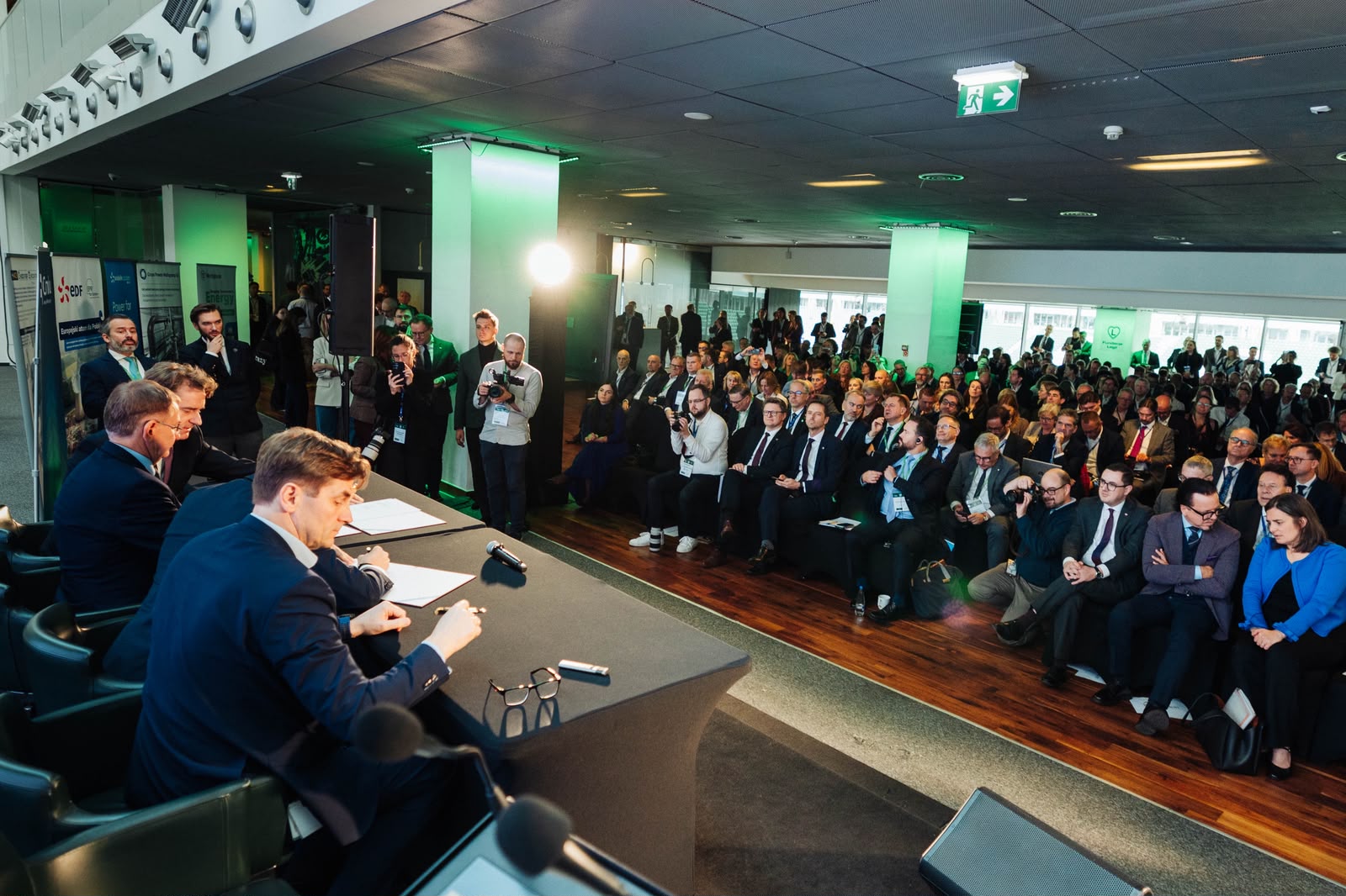 Mężczyźni w garniturach siedzą przy długim stole, podpisując dokumenty przed publicznością w nowoczesnej sali konferencyjnej. W tle roll-upy z logotypami firm i podświetlone filary.