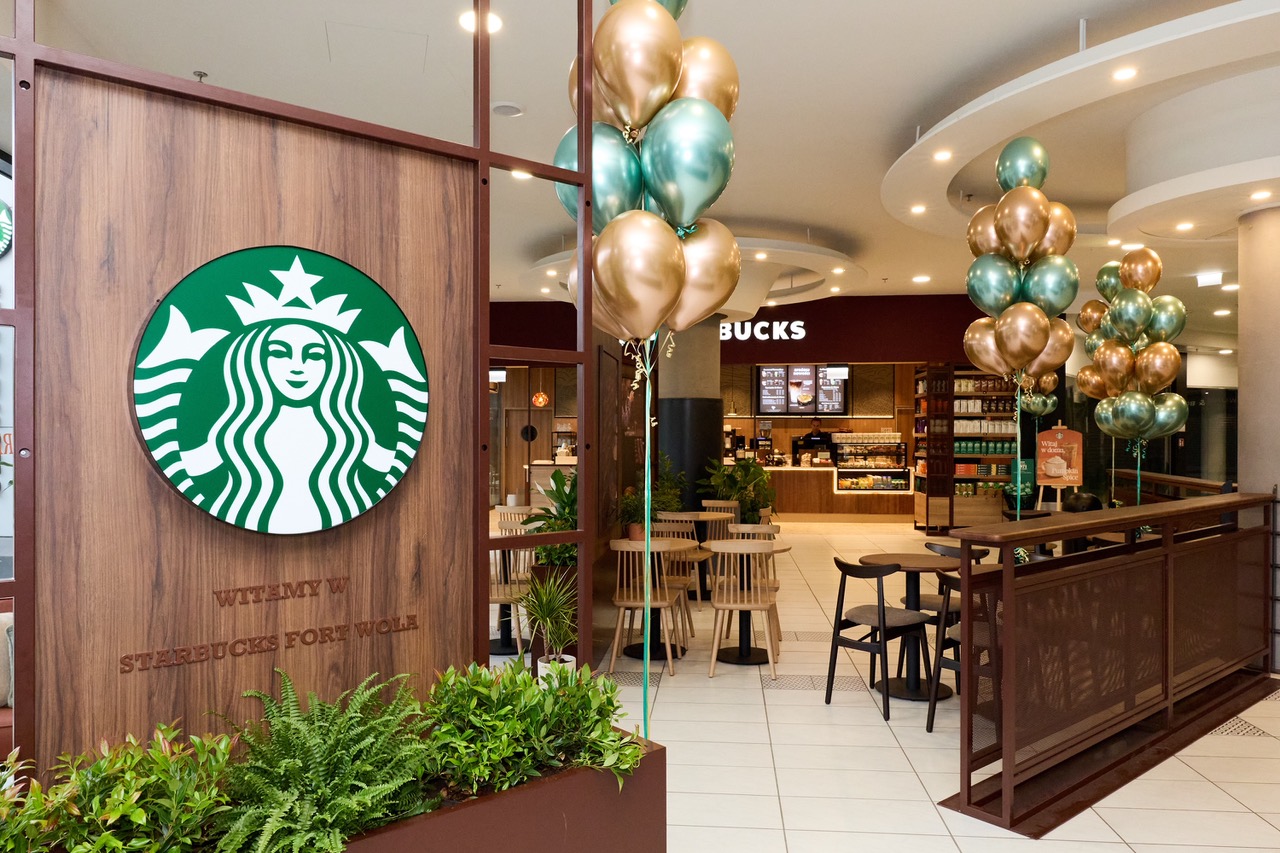Wejście do kawiarni Starbucks Fort Wola z okrągłym logo syreny na drewnianym panelu, jasne wnętrze z drewnianymi stolikami, czarnymi krzesłami, roślinami i balonami.