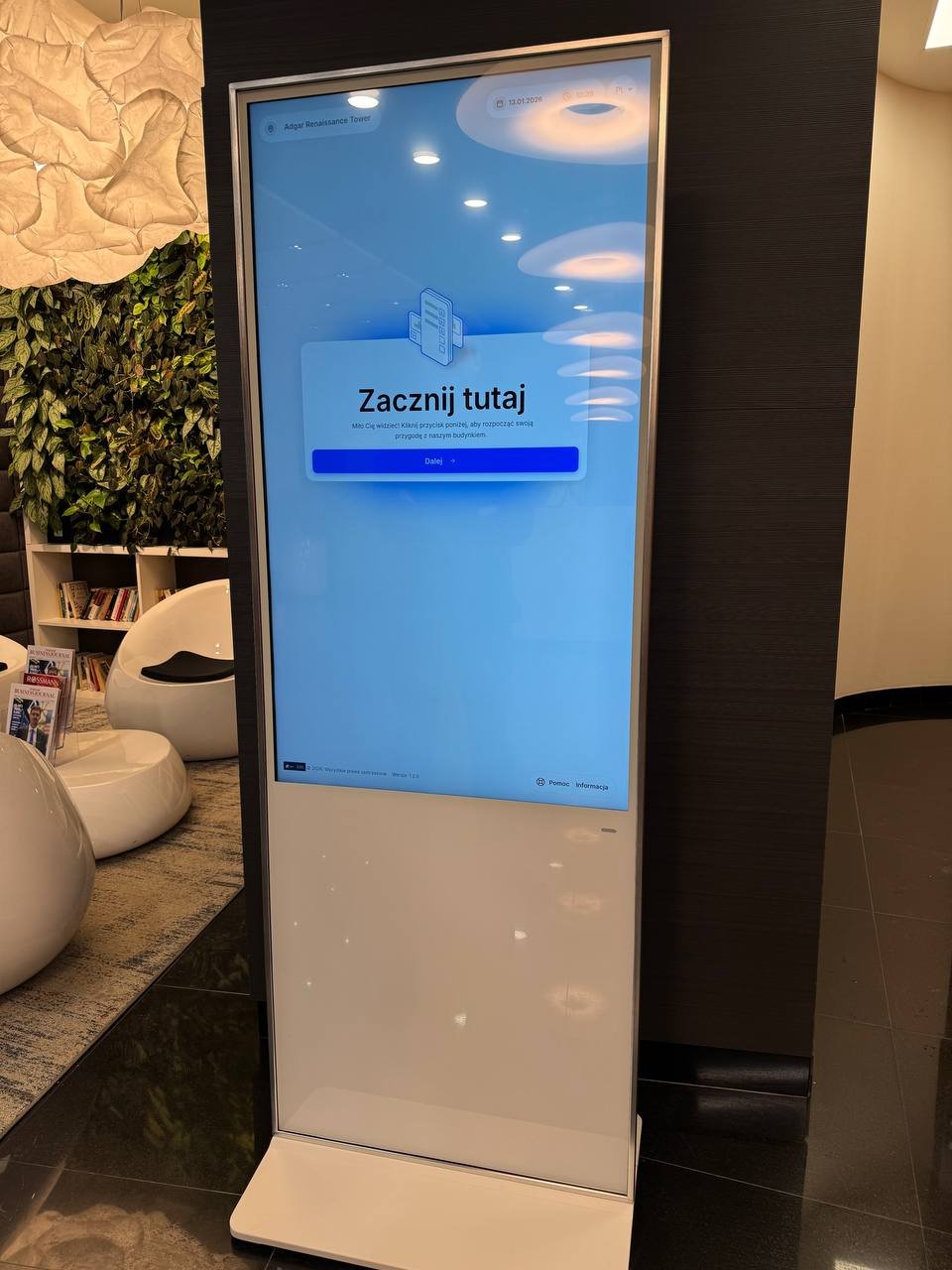 Pionowy, biały kiosk multimedialny z dużym ekranem dotykowym w nowoczesnym wnętrzu na tle ciemnej ściany. Na wyświetlaczu napis „Zacznij tutaj”.