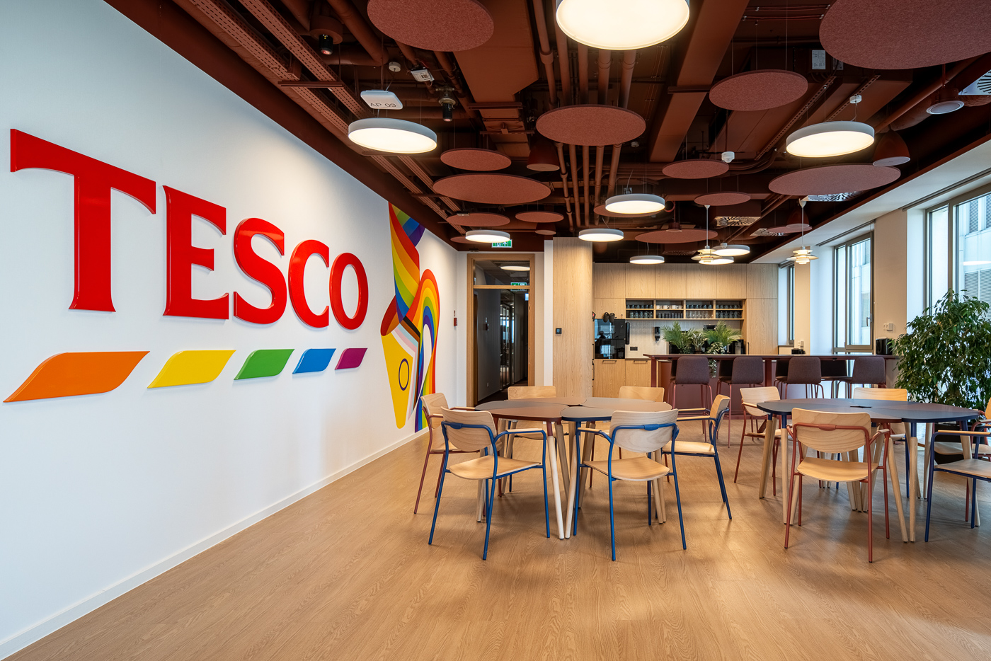 Nowoczesne wnętrze biurowe z drewnianymi stołami, aneksem kuchennym i ścianą z czerwonym napisem TESCO.