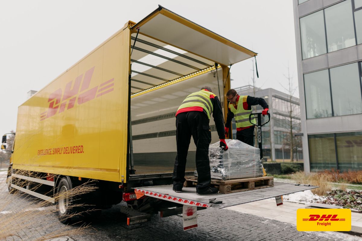 Żółta ciężarówka DHL z windą załadowczą oraz dwaj pracownicy rozładowujący paletę przed przeszklonym biurowcem.